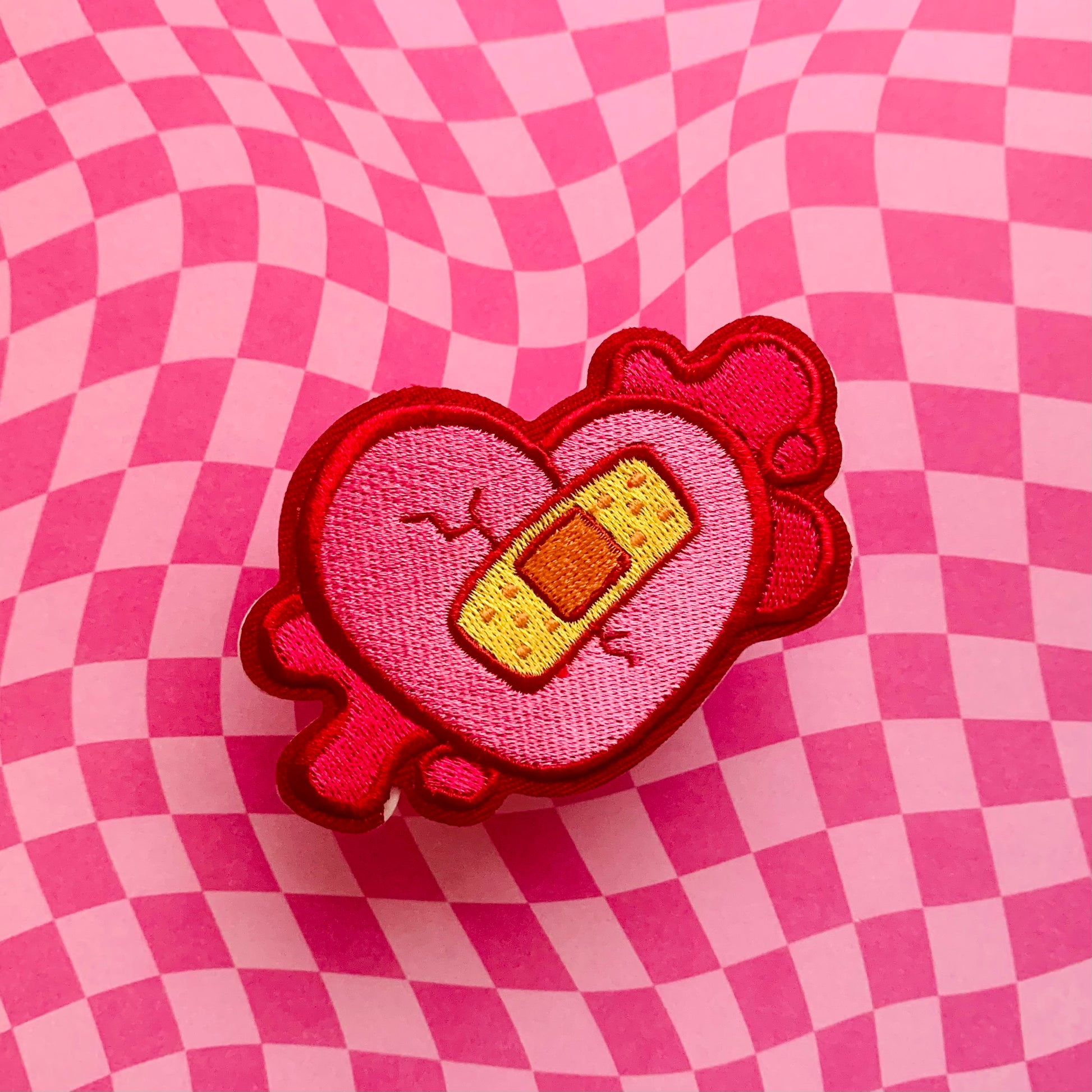 heart-shaped embroider patch with a bandage on a pink checkered background
